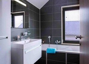 A modern bathroom with dark tiles, a white vanity, a large mirror, a bathtub, and a window with blinds. - Family Villa by the Lake (Froidchapelle)