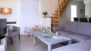 Una sala moderna con un sofá gris, una mesa de centro, una mesita con bebidas, una escalera de madera y un televisor sobre un mueble.
