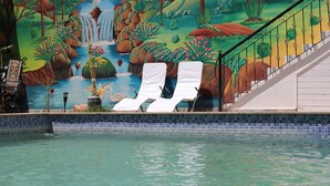 Une piscine agrémentée de deux chaises longues blanches et d’une fresque représentant une cascade et un paysage tropical.