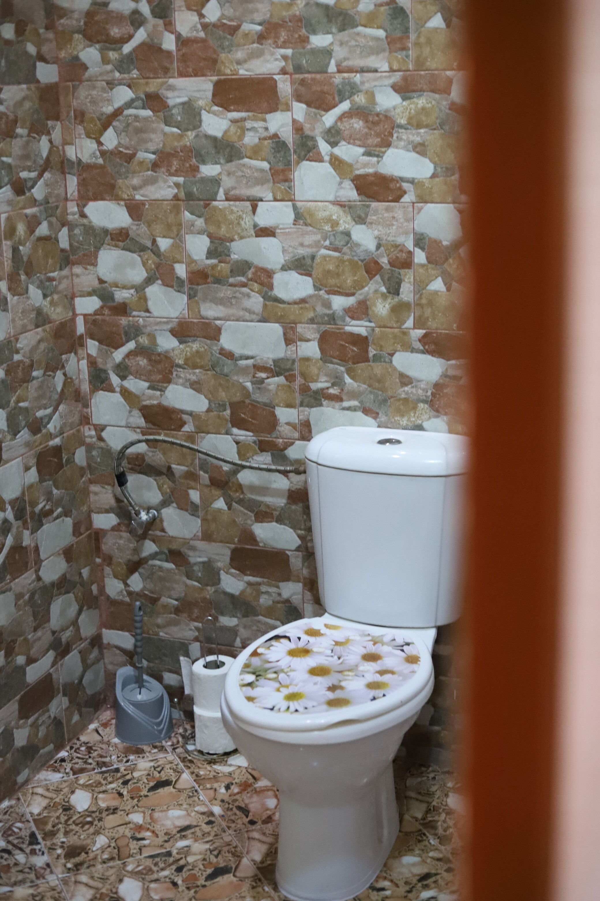 A bathroom with a mosaic tile wall, a toilet with a floral seat cover, and a plunger on the floor.