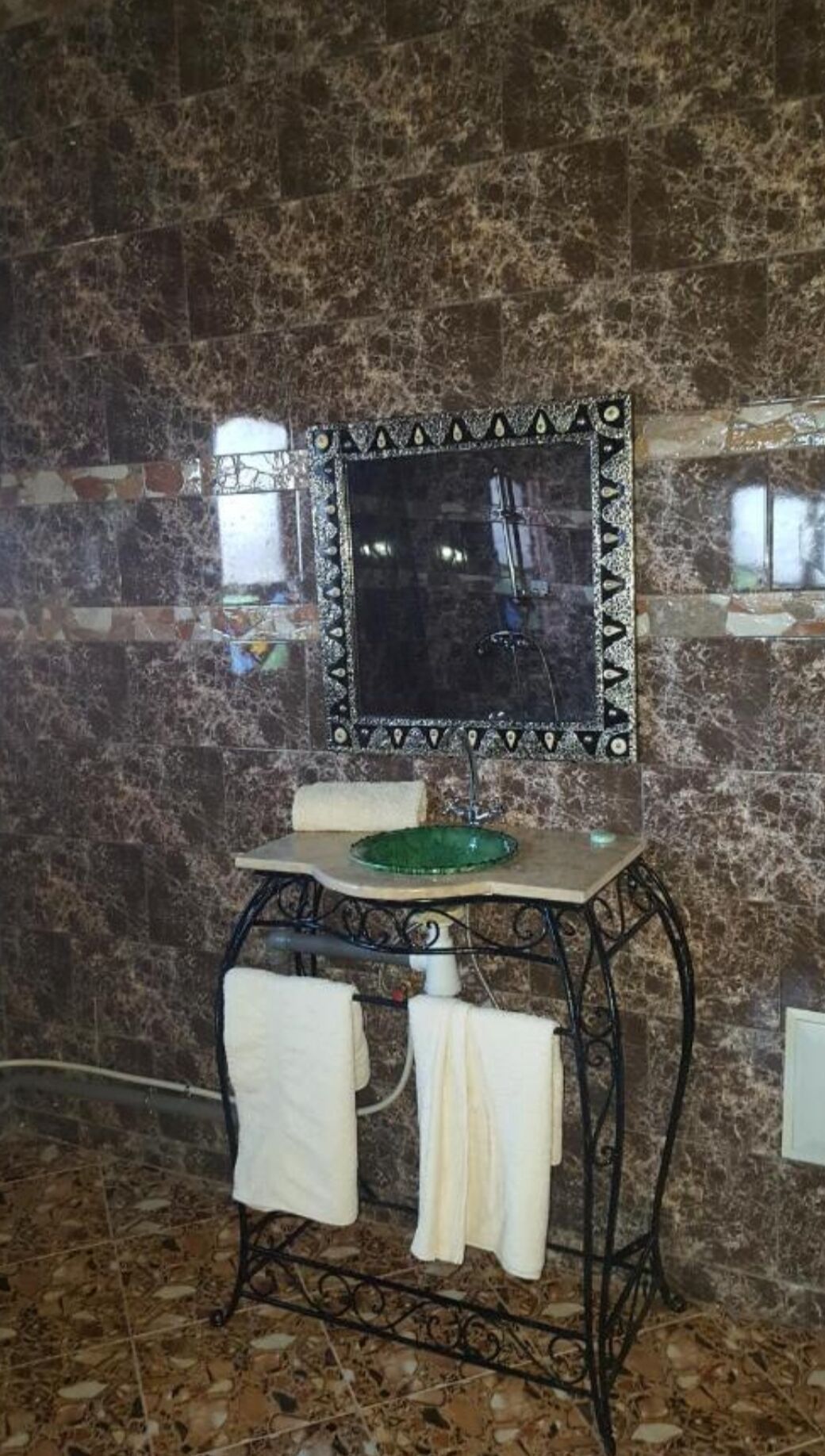 A bathroom with a marble wall, a sink with a green basin, and a mirror with a decorative frame.