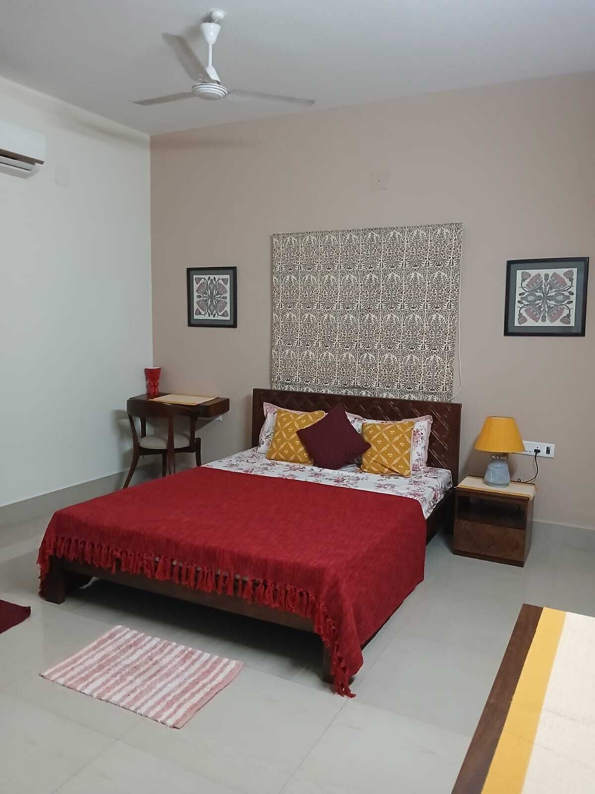 A bedroom with a bed, a nightstand, a chair, a red blanket, a patterned wall hanging, and a ceiling fan.