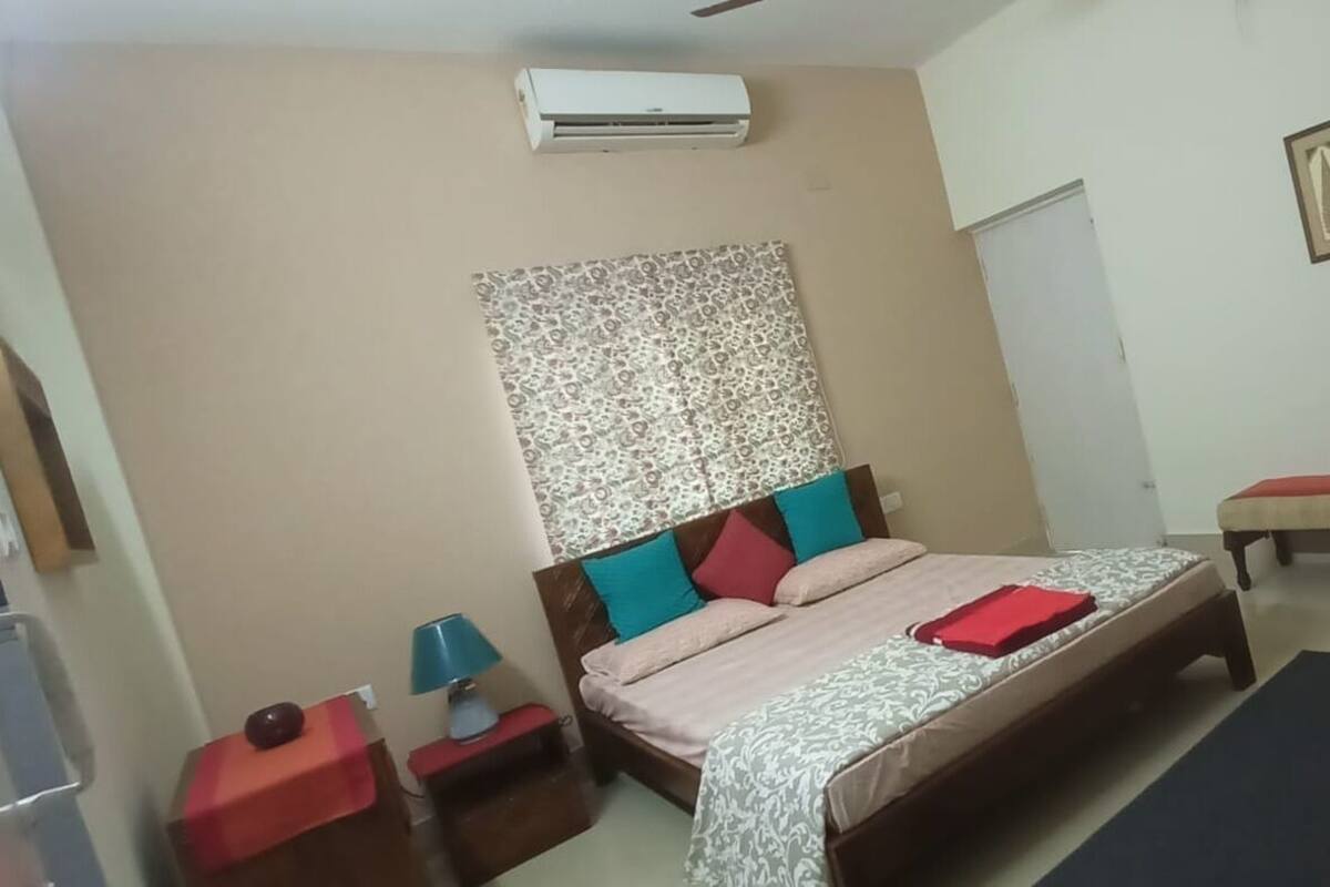 Een slaapkamer met een bed, nachtkastje, lamp, bankje en televisie.