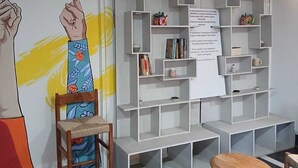 A room with a wall mural, a wooden chair, a small table, and a white shelving unit. - The Basic (Bhubaneshwar)