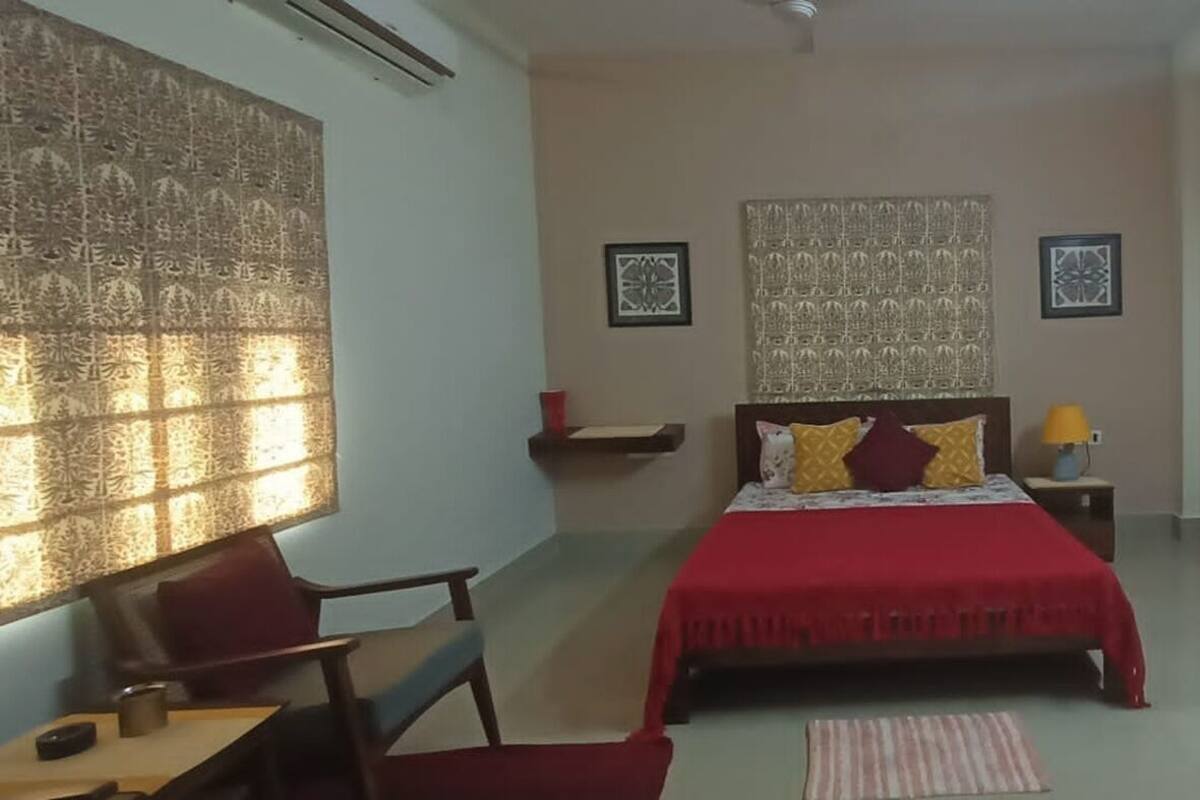 Een slaapkamer met een bed, een bureau, een stoel, een raam met gordijnen met patroon, en een muur met kunstwerken.