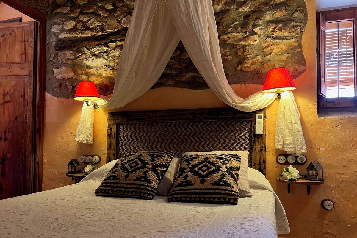 Un dormitorio con pared de piedra, cabecera de madera, cama con almohadas estampadas, dos lámparas de noche y una mesita con objetos decorativos.