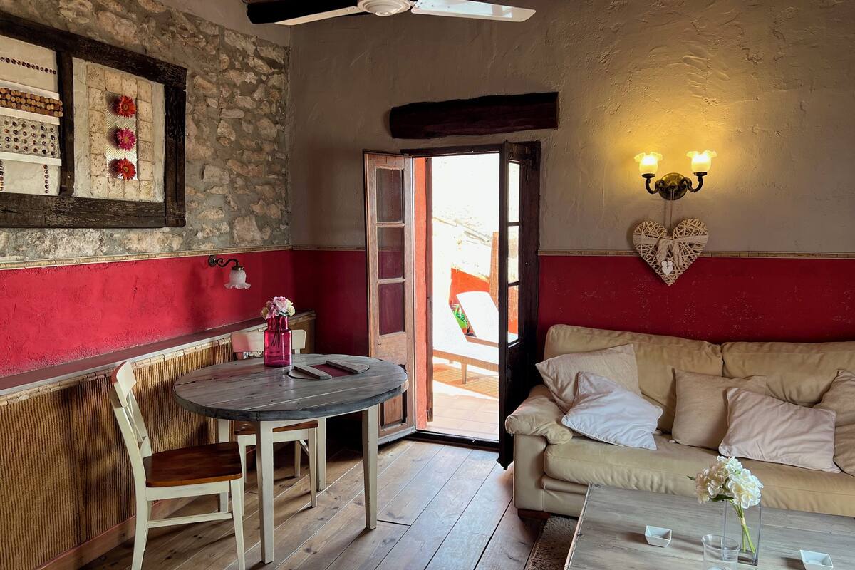 Una habitación acogedora con suelo de madera, una mesa redonda con sillas, un sofá, una lámpara fijada a la pared y una puerta que da al exterior.