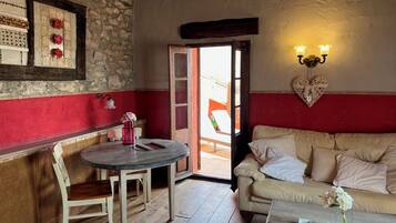 A cozy room with a wooden floor, a round table with chairs, a sofa, a wall-mounted light fixture, and a door leading outside.