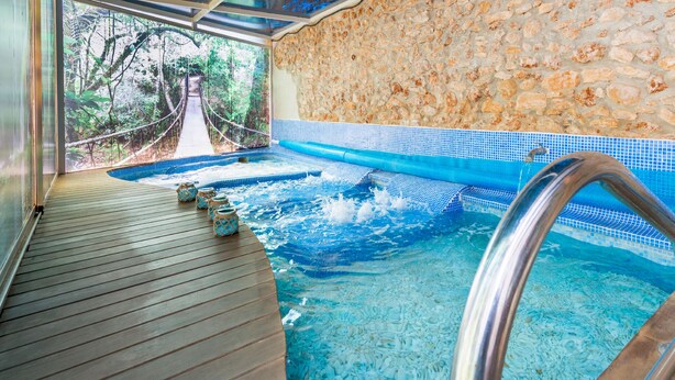 Un área de spa con terraza de madera, pared de piedra y una vista a un puente colgante al fondo.