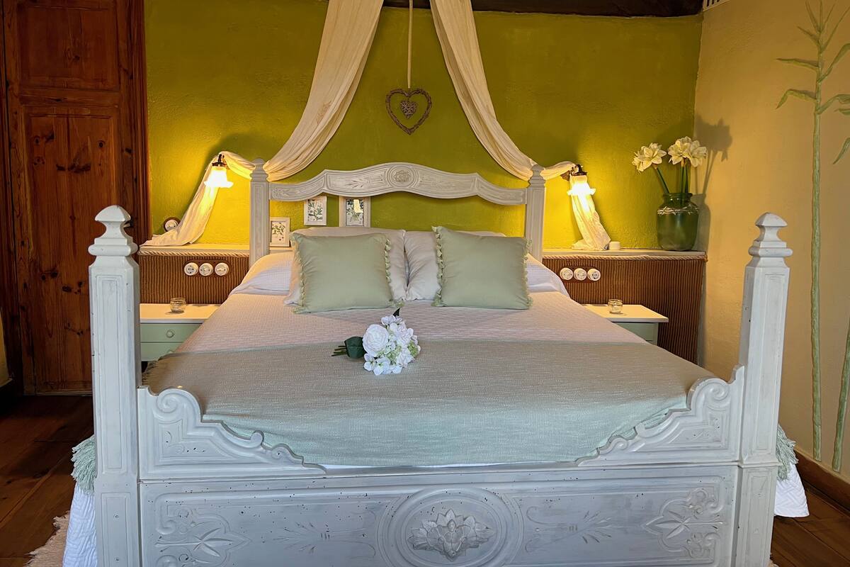 Una cama con dosel, dos mesitas de noche con lámparas, una decoración con forma de corazón y un florero con flores.