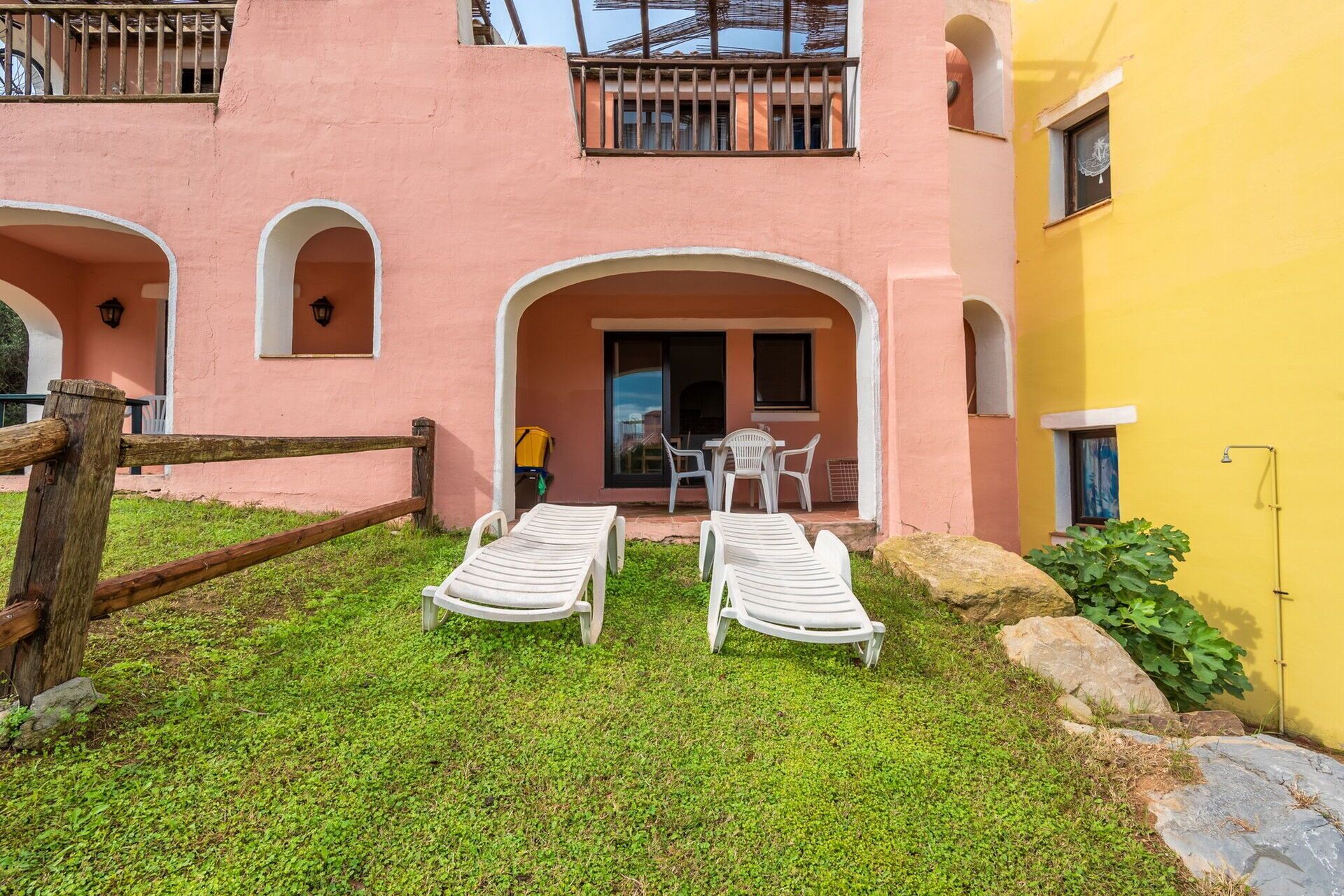 A building with a pink facade, a balcony, and a yellow wall. There are two white lounge chairs and a table with chairs outside.