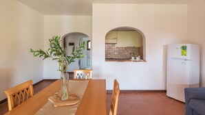 Un coin repas avec une table en bois, des chaises et un vase rempli de verdure. On aperçoit une cuisine à travers une arche, où se trouve un réfrigérateur.