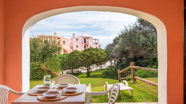 Un coin repas avec une table dressée pour quatre personnes, donnant sur un jardin et un bâtiment rose doté de nombreux balcons.