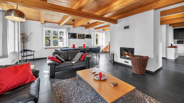 Una sala de estar moderna con chimenea, sofá de cuero negro, mesa de centro de madera y un comedor con cocina al fondo.