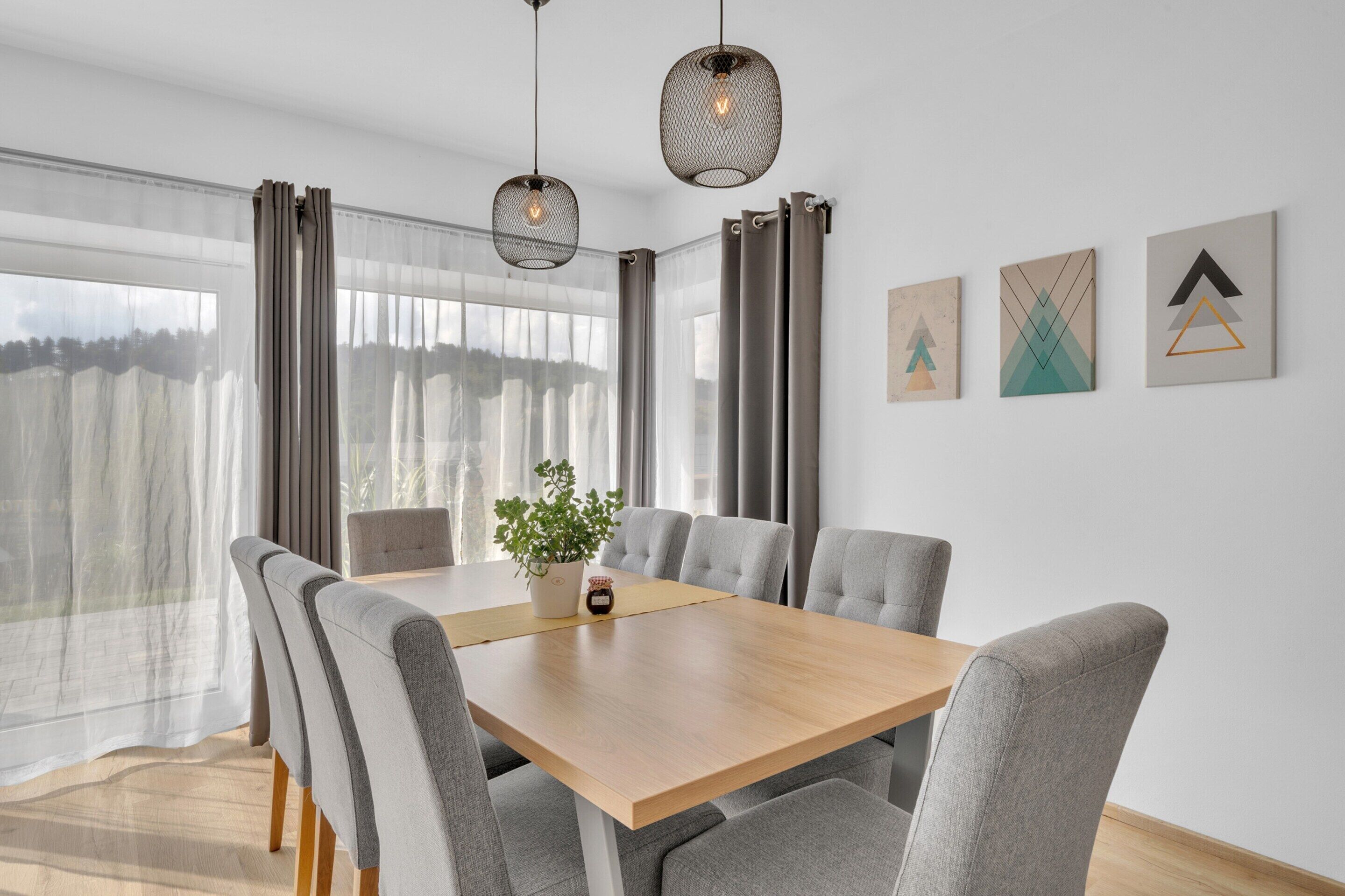 A modern dining area with a wooden table and grey upholstered chairs, large windows with sheer curtains, and geometric wall art.
