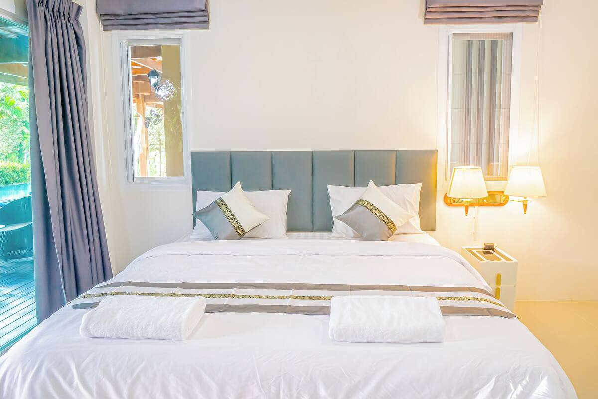 A neatly made bed with white linens and decorative pillows, a bedside lamp, and a window with curtains.