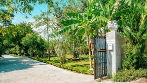 A paved pathway leading to a gated entrance with a statue, surrounded by lush greenery and banana trees. - Farinda Resort & Pool Villa (Chom Thong)