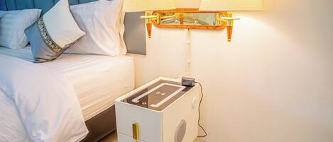 Una cama bien tendida con sábanas blancas y almohadones decorativos, una mesita de noche con una lámpara y un pequeño dispositivo electrónico, y un aplique en la pared.