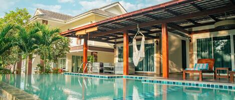 A swimming pool with a wooden deck and lounge chairs, surrounded by palm trees and a covered patio area.