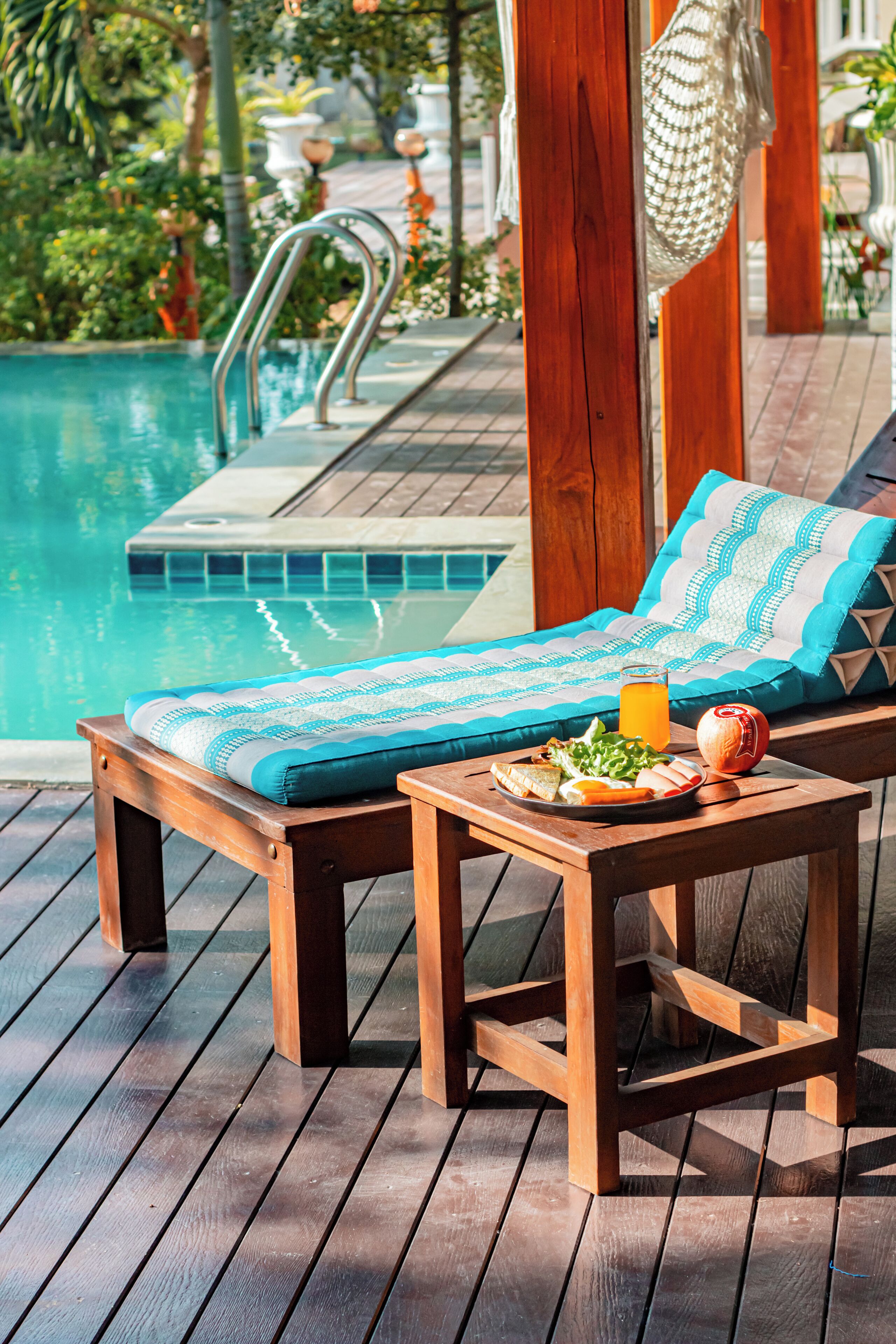 A wooden deck with a poolside lounge chair, a table with a drink and food, and a hanging net.
