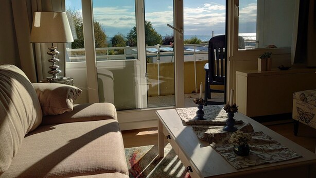 A living room with a sofa, a coffee table, and a view through a sliding glass door to an outdoor area with a chair and a table. - Holiday Home in Fehmarn by the Beach (Fehmarn)
