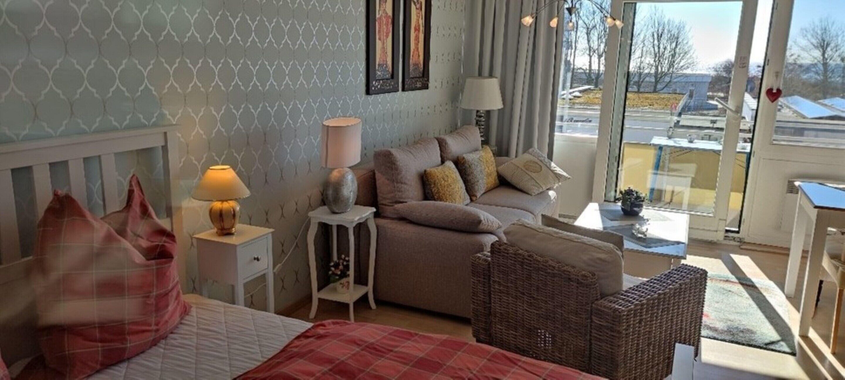 A bedroom with a bed, a nightstand, a lamp, a sofa, and a wicker chair.