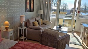 A bedroom with a bed, a nightstand, a lamp, a sofa, and a wicker chair. - Holiday Home in Fehmarn by the Beach (Fehmarn)