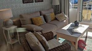 A living room with a sofa, cushions, a side table, a lamp, and a view through a glass door. - Holiday Home in Fehmarn by the Beach (Fehmarn)