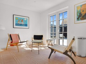 Una habitación con un ventanal grande, un cuadro enmarcado, una silla, un banco y un cubo de basura.