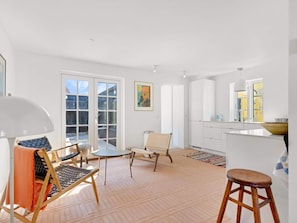 A spacious room with a large sliding glass door leading to a deck, a round table, two chairs, a wooden stool, and a kitchen area with cabinets and a window. - 5 Star Holiday Home in Lokken (Lokken)