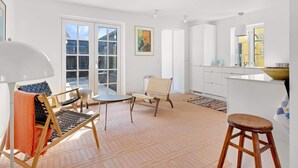 A spacious living area with a large sliding glass door, a round table, two chairs, a wooden stool, and a kitchen area with cabinets and a window. - 5 Star Holiday Home in Lokken (Lokken)