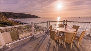 Un balcon avec une table ronde et des chaises en osier, donnant sur une étendue d’eau où le soleil se couche.