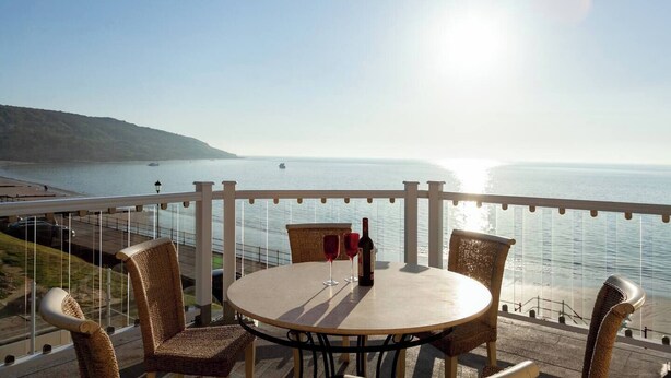 Une table ronde entourée de chaises en osier, sur un balcon donnant sur une étendue d’eau, avec une terre lointaine à l’horizon.