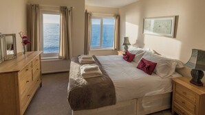 Une chambre avec un grand lit, une commode en bois, une table de chevet et une fenêtre donnant sur la mer.