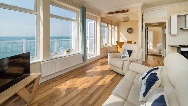A spacious living room with a large window overlooking the sea, a sofa, a TV, and a wooden floor. - Stunning Penthouse Apartment (Totland Bay)