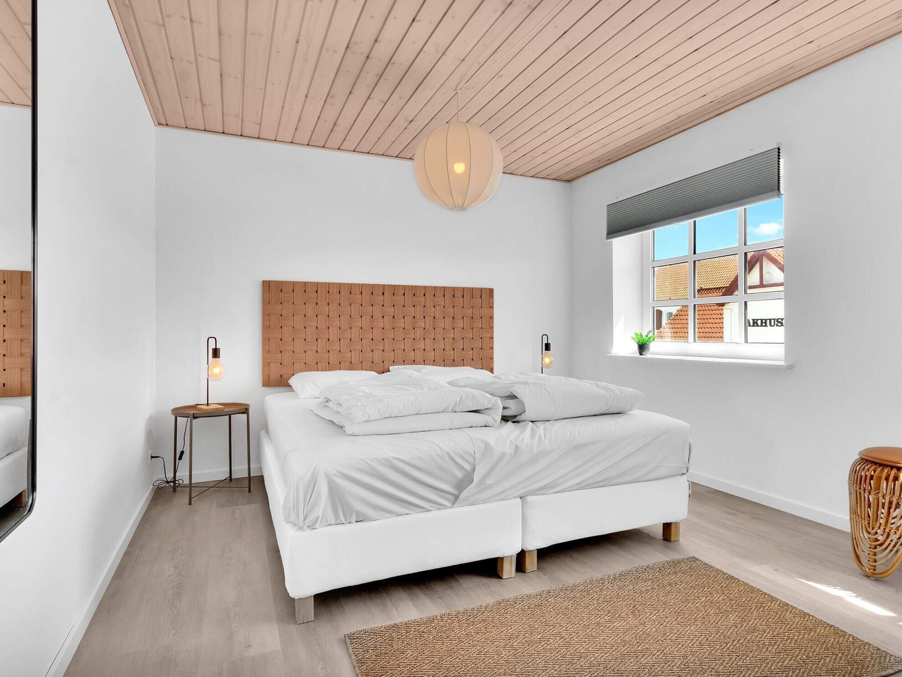 Un dormitorio moderno con techo de madera, una cama grande con ropa de cama blanca, dos lámparas de noche, una alfombra tejida y una ventana con vistas a edificios.