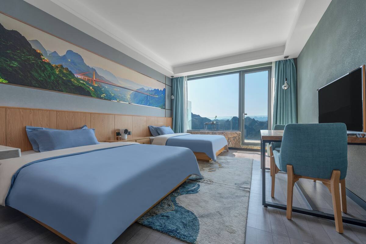 Ein Hotelzimmer mit zwei Betten, einem Schreibtisch, einem Stuhl, einem großen Fenster und einem Bild einer Brücke.