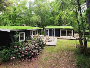 A cabin with a green roof, a deck, and a garden with pink flowers. - 4 Person Holiday Home in Frederiksvaerk (Frederiksvaerk)