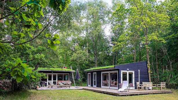 A modern cabin with a green roof, surrounded by a lush forest. It features a deck with outdoor seating and a covered patio area. - 4 Person Holiday Home in Frederiksvaerk (Frederiksvaerk)