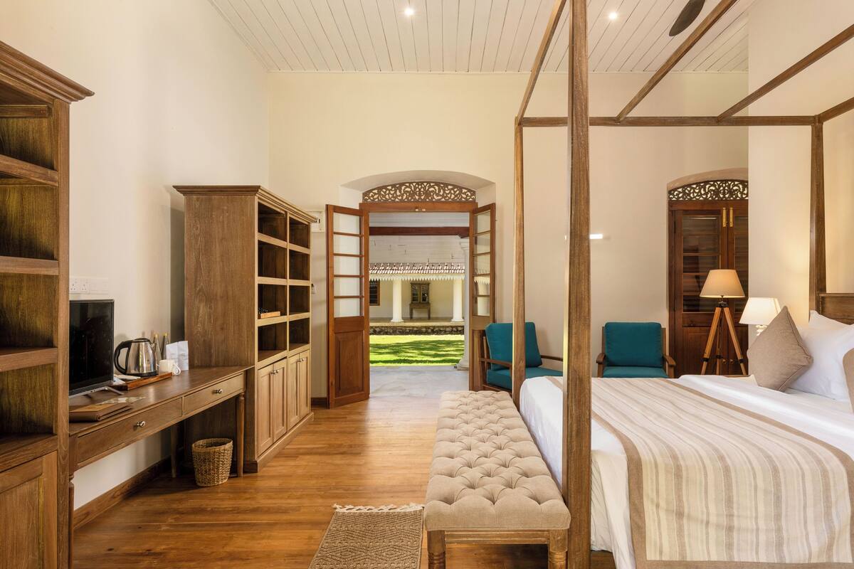 A bedroom with a four-poster bed, wooden furniture, a desk with a computer, and a view of a garden.