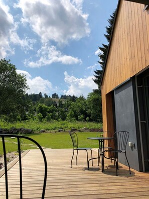 Eine Holzterrasse mit Metallstühlen und einem Tisch, ein Gebäude mit geneigtem Dach und eine grüne Landschaft mit Bäumen und blauem Himmel.