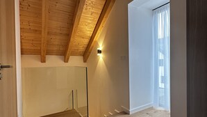 Ein geräumiges Zimmer mit Holzdielen, einer schrägen Decke mit sichtbaren Balken, einer Glaskonstruktion als Treppe und einem großen Fenster mit leichten Vorhängen.