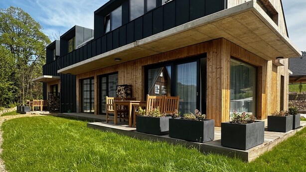 Ein modernes, zweistöckiges Haus mit überdachter Terrasse, großen Fenstern und einem gepflegten Rasen.