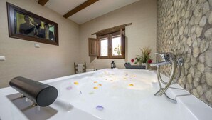 Une salle de bain avec une baignoire remplie de mousse et de pétales de rose, une fenêtre à cadre en bois et un mur en pierre.