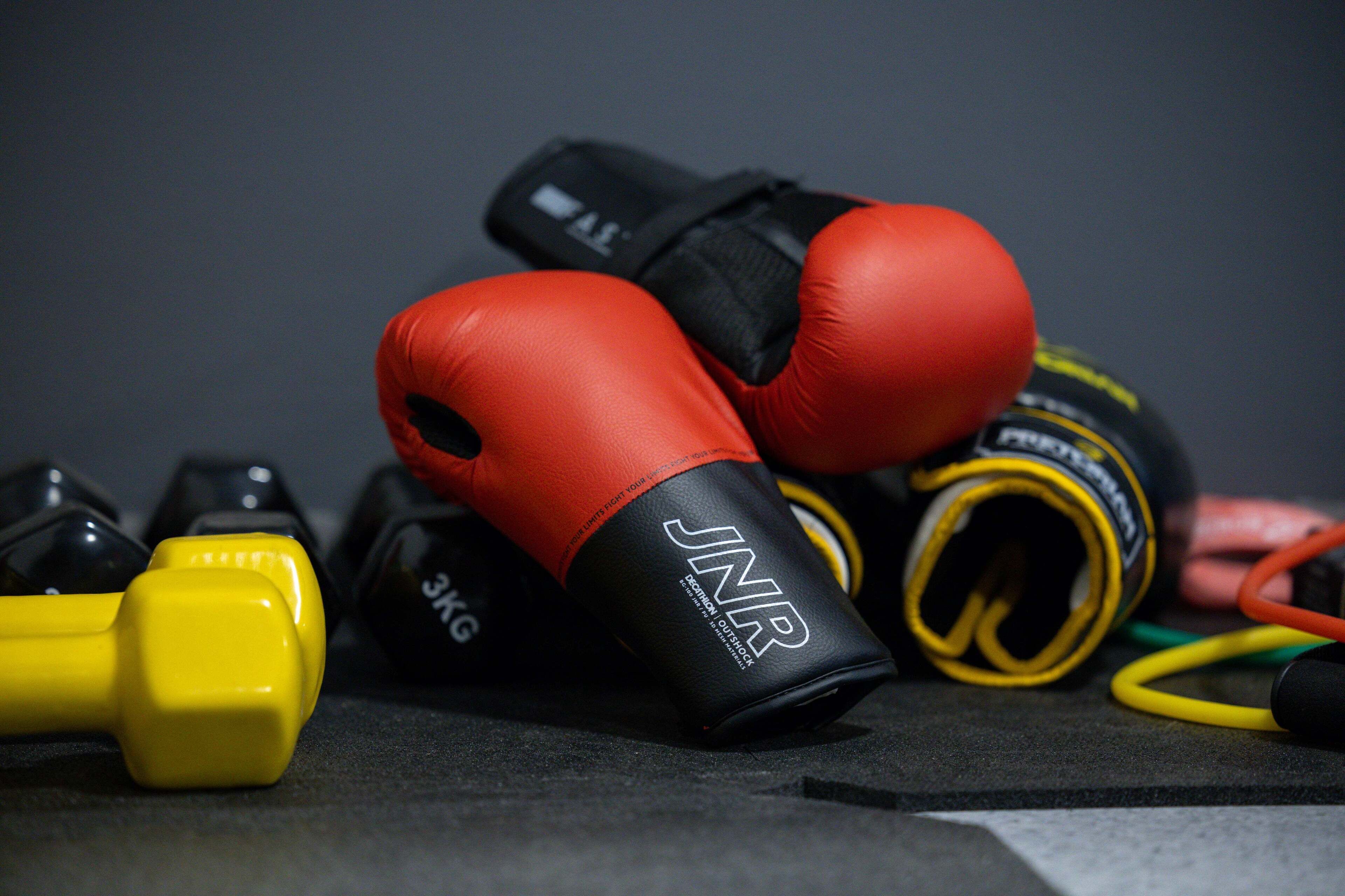 A pair of red boxing gloves, a yellow dumbbell, and a black and yellow resistance band on a dark surface.