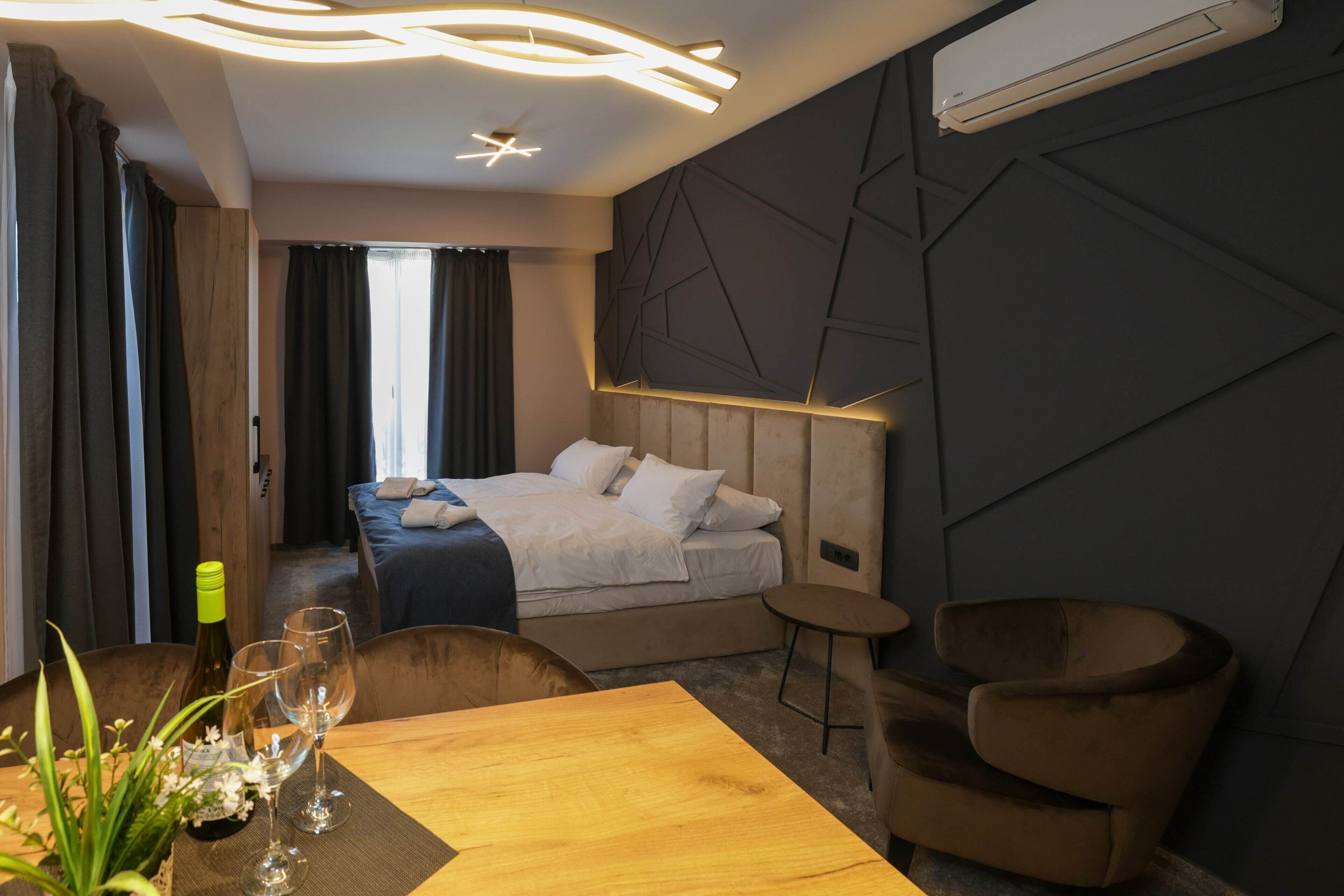 A modern hotel room with a bed, a dining table with chairs, a small side table, and a potted plant.