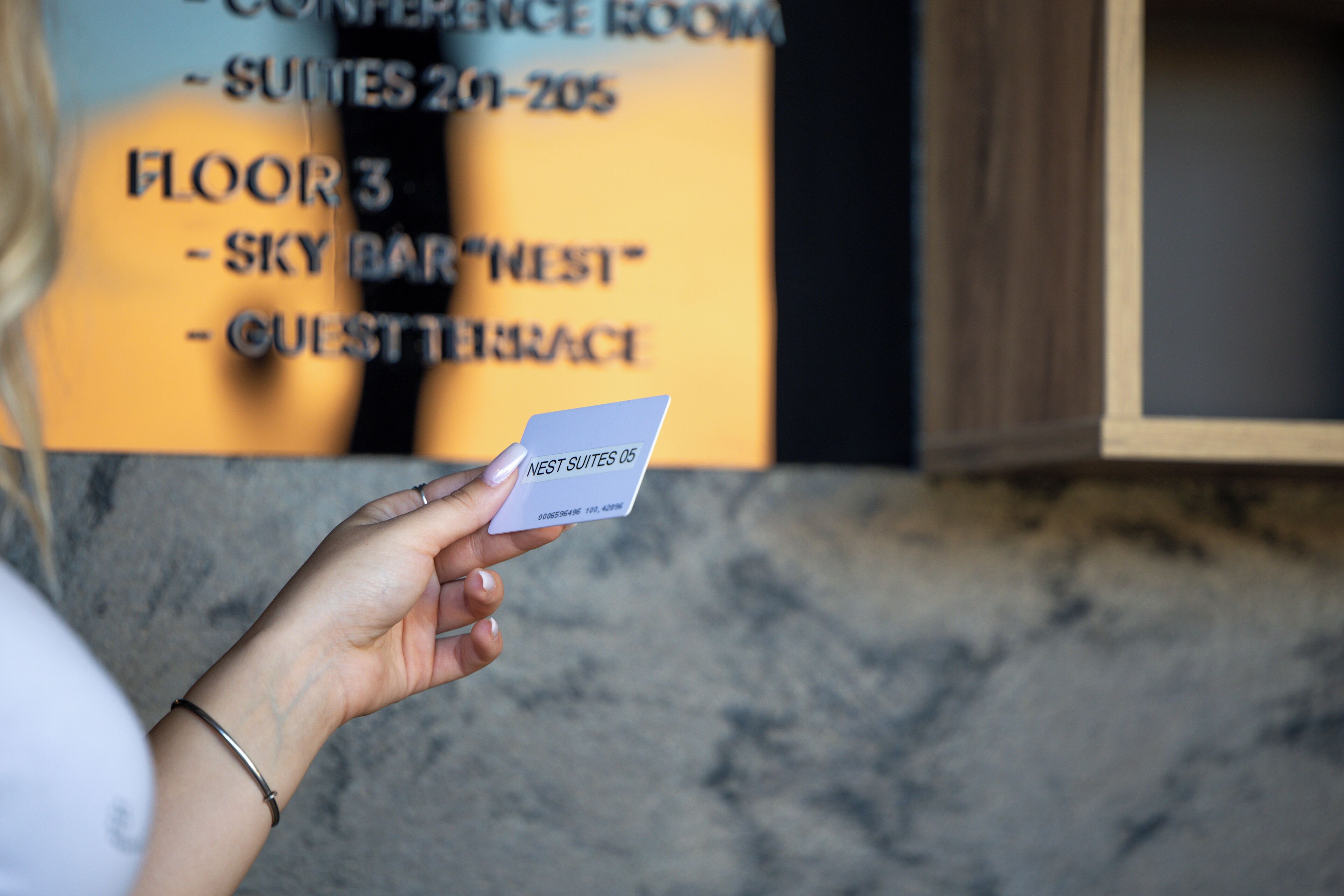 A hand holding a card with "NEST SUITES 05" printed on it, in front of a sign listing "FLOOR 3 - SKY BAR" and "GUEST BRACE".