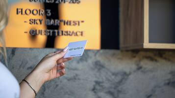 A hand holding a card with "NEST SUITES 05" printed on it, in front of a sign listing "FLOOR 3 - SKY BAR" and "GUEST BRACE".