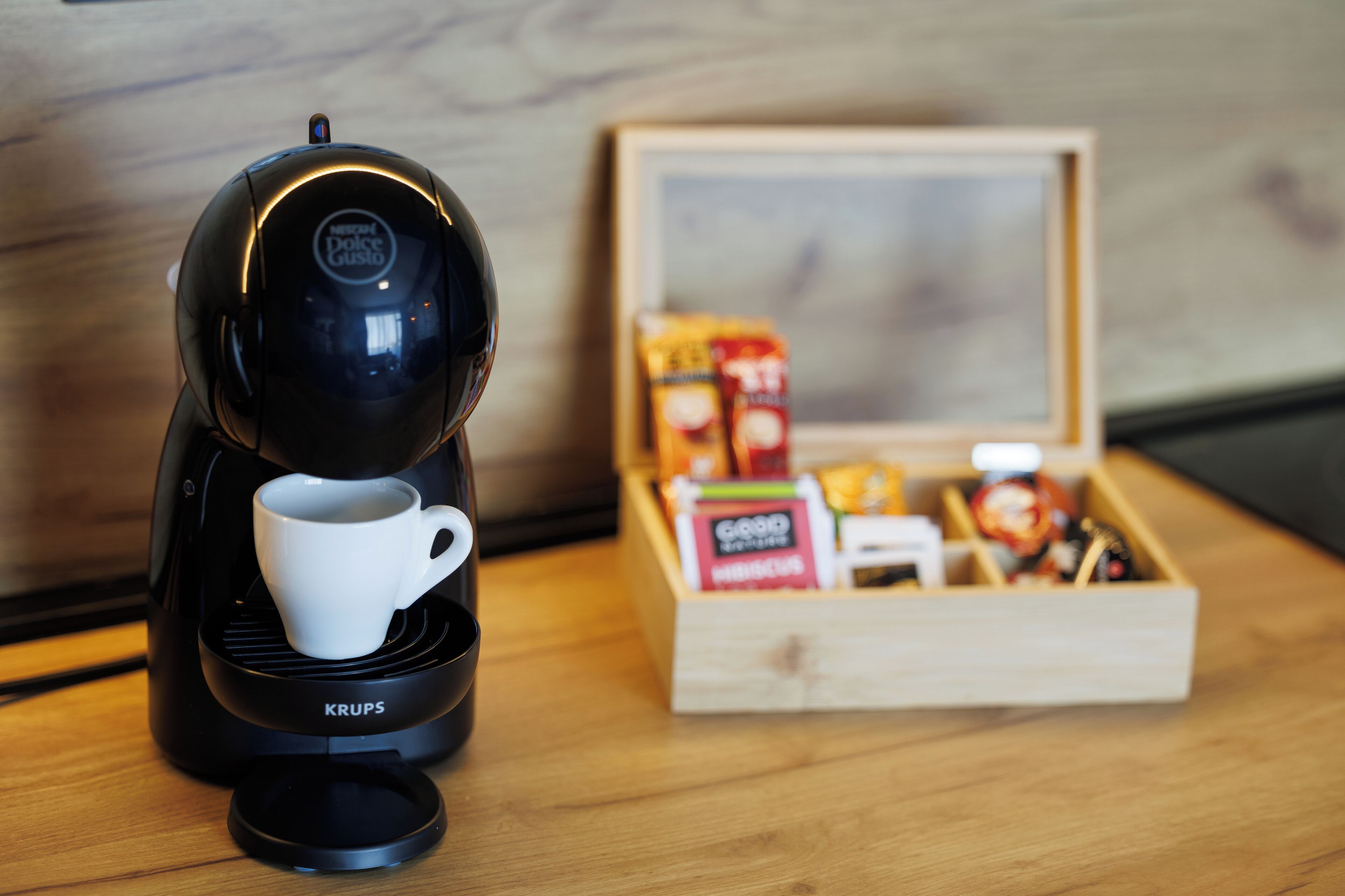 A Krups coffee maker with a cup, a wooden box of coffee pods, and a box of milk on a wooden surface.