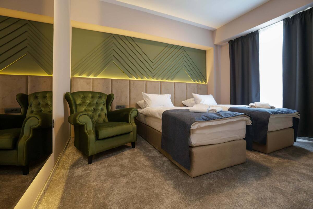 A hotel room with a large bed, two green armchairs, a headboard with geometric patterns, and a view of the outdoors through a window.